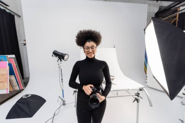 Siyah balıkçı yaka kazaklı ve gözlüklü kaygısız Afrikalı Amerikalı fotoğrafçı modern fotoğraf stüdyosundaki spot ışıklarının yanındaki kameraya bakıyor.