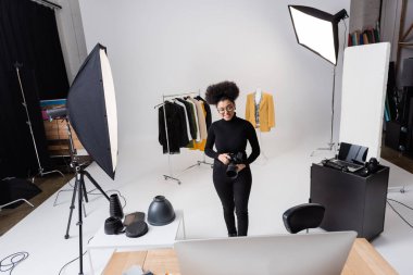 Mutlu Afro-Amerikan içerik yapımcısı fotoğraf stüdyosunda moda kıyafetlerin yanında dijital kamera ve ışıklandırma ekipmanı ile