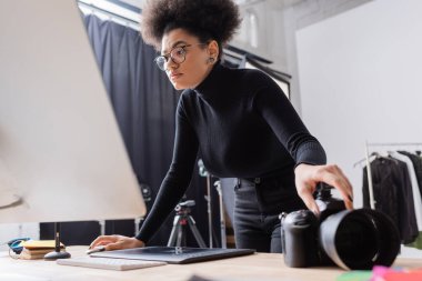 Afro-Amerikan içerik üreticisi fotoğraf stüdyosundaki grafik tablet ve dijital kameranın yanındaki bilgisayar monitörüne bakıyor.