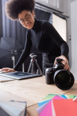 blurred african american content maker touching digital camera near graphic tablet in photo studio clipart