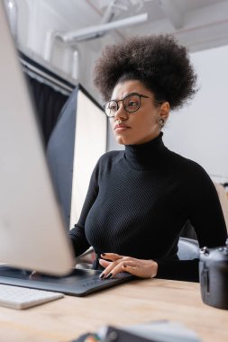 Siyah balıkçı yaka kazak ve gözlüklü konsantre Afro-Amerikan rötuş cihazı fotoğraf stüdyosunda bilgisayar ve grafik tablet üzerinde çalışıyor.