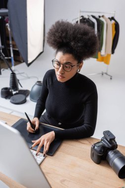 Gözlüklü Afro-Amerikan rötuşçusunun dijital fotoğraf stüdyosundaki fotoğraf makinesinin yanında grafik tablet ve bilgisayar üzerinde çalışırken yüksek açılı görüntüsü.