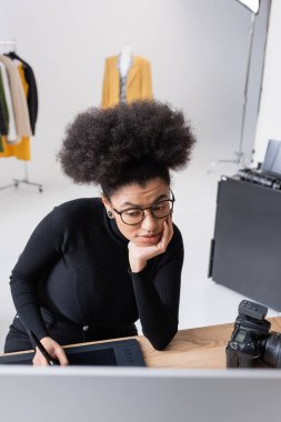 Gözlüklü düşünceli Afro-Amerikan rötuş cihazı fotoğraf stüdyosunda grafik tablet üzerinde çalışırken bilgisayar ekranına bakıyor.