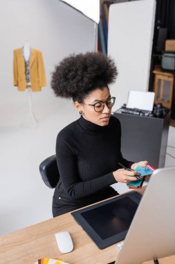 Afro-Amerikan rötuş cihazı fotoğraf stüdyosunda bilgisayar monitörü ve grafik tabletinin yanında renk örnekleri tutuyor.