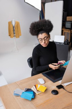Gözlüklü Afro-Amerikan rötuş cihazı, fotoğraf stüdyosunda grafik tabletinin yanında renk örnekleri ve bilgisayar monitörü tutuyor.