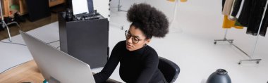 Gözlüklü ve siyah balıkçı yakalı Afro-Amerikan içerik müdürünün fotoğraf stüdyosunda bilgisayar üzerinde çalışmasının yüksek açılı görüntüsü.
