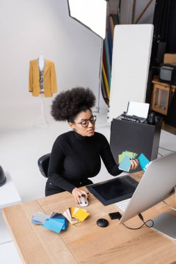 Fotoğraf stüdyosunda bilgisayar ve grafik tablet üzerinde çalışırken elinde renk örnekleri tutan Afrikalı Amerikan rötuşçusunun yüksek açılı görüntüsü
