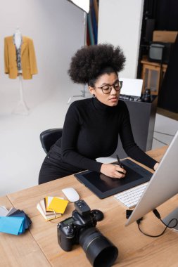 Gözlüklü Afro-Amerikan rötuş cihazı dijital kamera ve fotoğraf stüdyosundaki renk örnekleri yanında grafik tablet ve bilgisayar üzerinde çalışıyor.