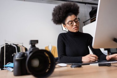 Afro-Amerikan rötuşçusu bilgisayarın yanında grafik tablet kullanıyor ve fotoğraf stüdyosunda bulanık dijital kamera kullanıyor.