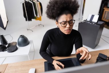 Düşünceli Afrikalı Amerikalı rötuşçu fotoğraf stüdyosunda grafik tablet üzerinde çalışırken bulanık bilgisayar monitörünü işaret ediyor.