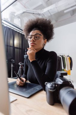 Siyahi balıkçı yaka kazak ve gözlük takmış, fotoğraf stüdyosundaki bulanık ekranın yanında grafik tablet üzerinde çalışan düşünceli Afro-Amerikan rötuş cihazı.