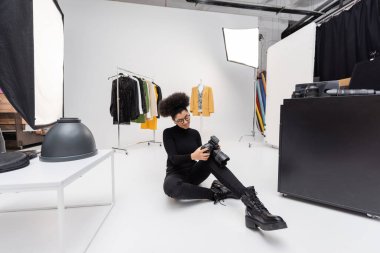 Gülümseyen Afrikalı Amerikalı fotoğrafçı fotoğraf stüdyosundaki moda kıyafetlerin ve projektörlerin yanında otururken dijital kameraya bakıyor.