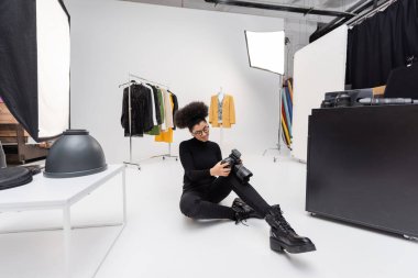 smiling african american photographer looking at digital camera while sitting near trendy clothes and floodlights in photo studio clipart
