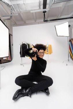 Mutlu Afro-Amerikalı kadın fotoğraf stüdyosunda moda kıyafetlerin ve spot ışıklarının yanında otururken dijital kamerada fotoğraf çekiyor.