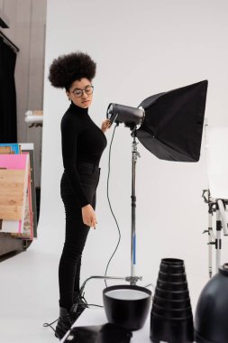 Gözlüklü ve siyah elbiseli Afro-Amerikan yapımcı fotoğraf stüdyosundaki ışıklandırma ekipmanlarına bakıyor.