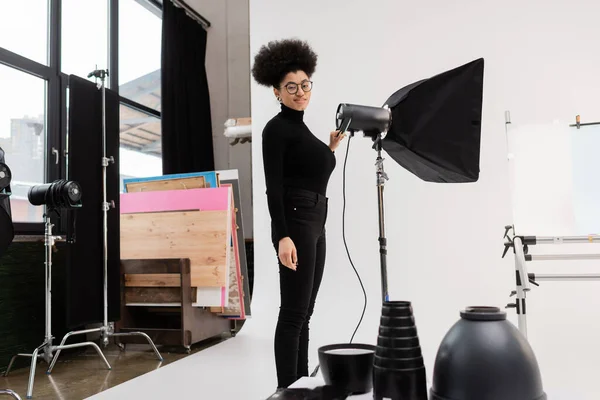 アフリカ系アメリカ人の眼鏡と黒い服のコンテンツマネージャーが写真スタジオで反射板の近くで笑っています — ストック写真