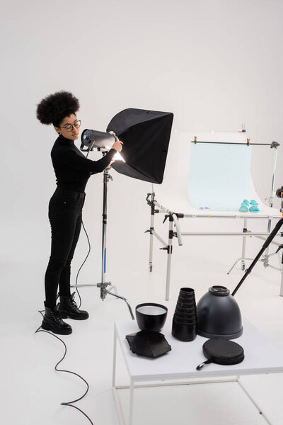 Африканский производитель контента, собирающий прожектор рядом со столом для съемок и осветительным оборудованием в фотостудии 