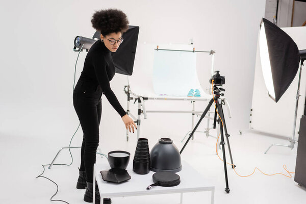 стильный африканский производитель контента достигает осветительного оборудования рядом с рефлектором Softbox и цифровой камерой на штативе в фотостудии