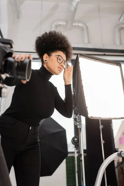stylish african american content manager adjusting eyeglasses near blurred digital camera and softbox reflector in photo studio