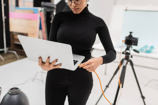 african american content producer connecting cable to laptop near blurred digital camera in photo studio