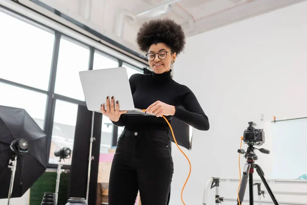 cheerful african american content manager plugging wire in laptop near tripod with digital camera in modern photo studio
