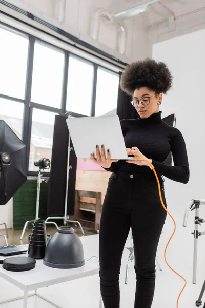 african american content manager in eyeglasses using laptop while standing near lighting equipment in photo studio