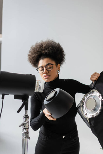 african american content manager in eyeglasses holding reflector while assembling strobe lamp in photo studio