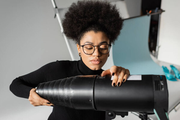 african american content producer in black turtleneck and eyeglasses assembling strobe spotlight in photo studio