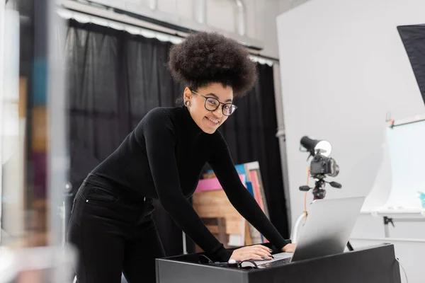 stylish and joyful african american content maker looking at camera and using laptop in photo studio