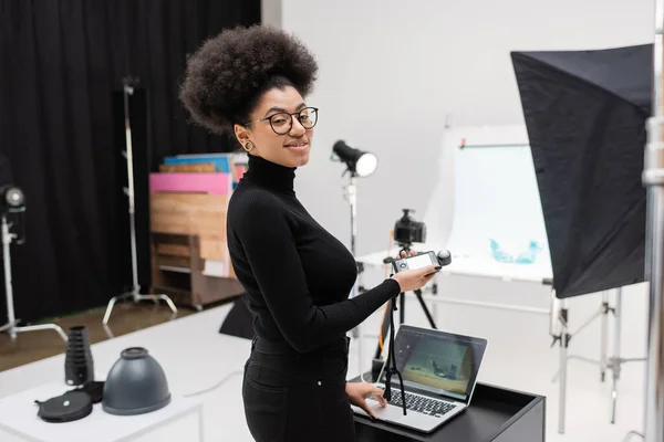 stock image happy african american content maker holding exposure meter and looking at camera near laptop in photo studio