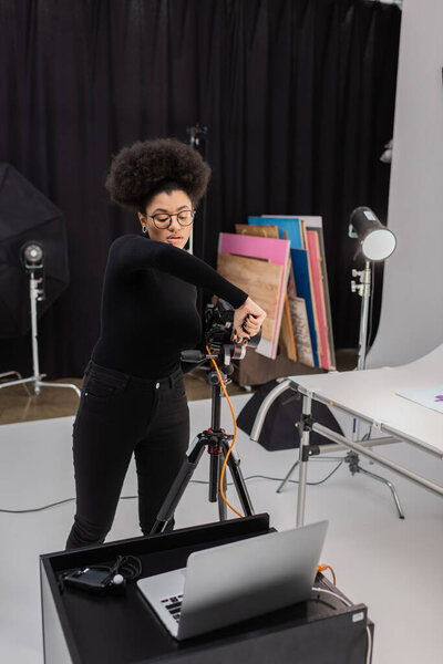 african american content producer in eyeglasses assembling digital camera on tripod near laptop in photo studio