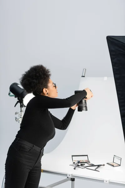 african american content manager taking photo of beauty tools and decorative cosmetics on shooting table in photo studio
