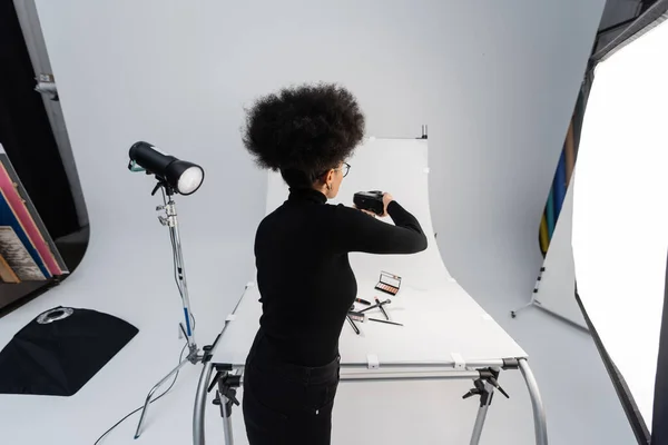back view of african american photographer taking picture of beauty tools and decorative cosmetics on shooting table in photo studio