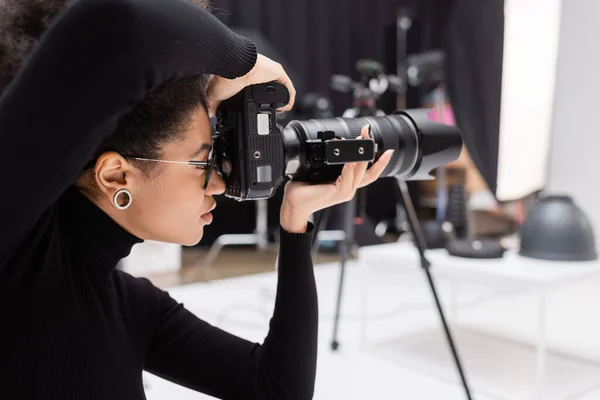 非洲裔美国摄影师穿着黑色高领毛衣在摄影棚的专业数码相机上拍照的侧视图 — 图库照片
