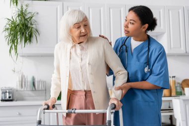 brunette multiracial nurse in blue uniform supporting senior woman with grey hair walking with help of walker at home  clipart