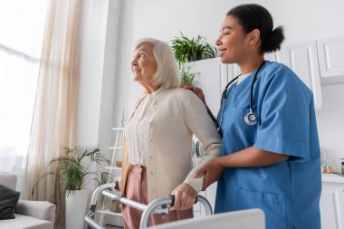cheerful multiracial nurse in blue uniform supporting happy senior woman with grey hair walking with help of walker at home  clipart