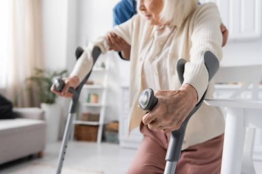 cropped view of multiracial caregiver in uniform helping senior woman using crutches to walk at home  clipart