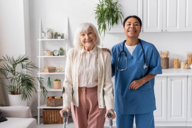 positive retired woman with grey hair using crutches while walking near happy multiracial nurse at home  clipart