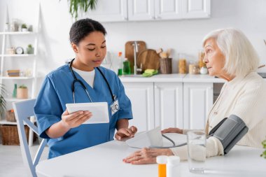 Sayısal tablet kullanan çok ırklı hemşire ve gri saçlı yaşlı kadının tansiyonunu ölçüyor. 