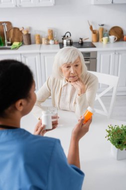 Esmer, çok ırklı hemşirenin elinde ilaç tutarken gri saçlı, emekli bir kadınla konuşmasının genel görüntüsü.