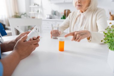 cropped view of senior woman pointing at bottle with medication near multiracial nurse on blurred foreground  clipart