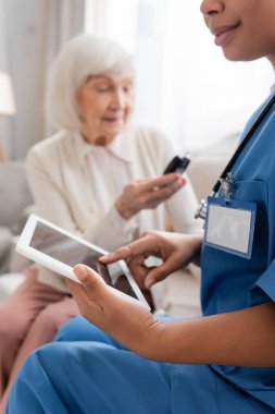 Çok ırklı hemşire bulanık arkaplanda yaşlı bir kadının yanında boş ekranlı dijital tablet kullanıyor. 