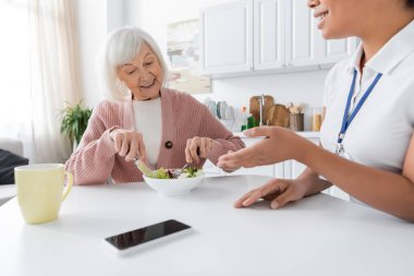 Gri saçlı, neşeli, çok ırklı bakıcının yanında öğle yemeği yiyen mutlu son sınıf öğrencisi kadın. 