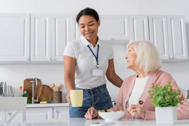 Mutfakta yemek yiyen, gri saçlı, emekli bir kadının yanında çay içen çok kültürlü sosyal hizmet görevlisi. 