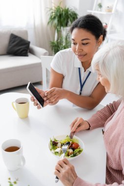 Mutfakta öğle yemeği yiyen yaşlı bir kadının yanındaki akıllı telefonu işaret eden mutlu sosyal hizmet görevlisi. 