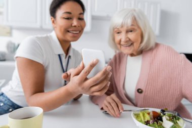 Mutfakta öğle yemeği sırasında üst düzey kadınlara akıllı telefon gösteren çok ırklı bir sosyal hizmet görevlisi. 