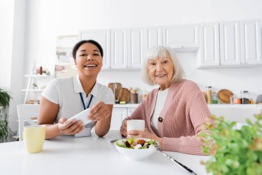 Mutfakta emekli bir kadınla yemek yerken elinde akıllı telefon tutan mutlu çok ırklı sosyal hizmet görevlisi. 