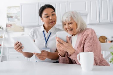 Esmer, çok ırklı sosyal hizmetler görevlisi elinde dijital tabletle mutfakta akıllı telefonlu yaşlı bir kadının yanında. 