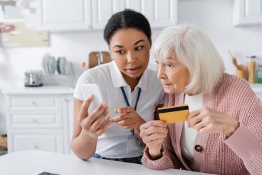 Esmer, çok ırklı sosyal hizmetler görevlisi kredi kartı olan yaşlı bir kadının yanında akıllı telefon tutuyor. 
