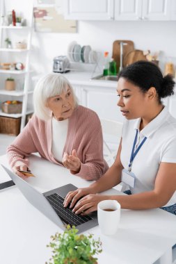 Çok uluslu sosyal hizmetler görevlisi kredi kartı olan yaşlı bir kadının yanında dizüstü bilgisayar kullanıyor. 